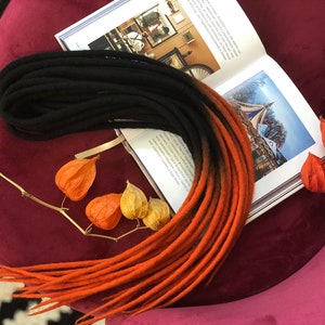 May include: A set of black and orange ombre wool dreadlocks. The dreadlocks are arranged in a circle on a burgundy surface.
