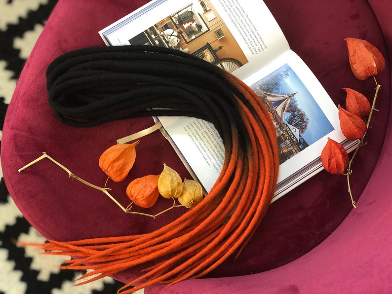 May include: A set of black and orange ombre wool dreadlocks. The dreadlocks are arranged in a circle on a burgundy surface.