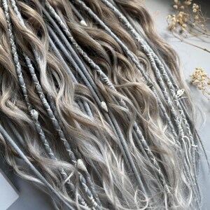 May include: Close-up of a set of gray and light brown dreadlocks. The dreadlocks are adorned with white thread and small decorative charms. The hair has a wavy texture and is arranged in a cascading pattern.