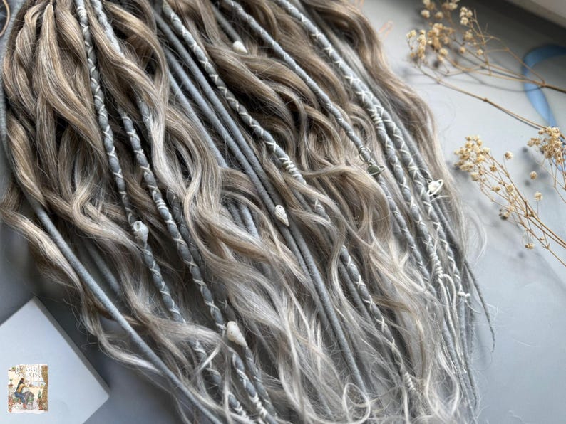 May include: Close-up of a set of gray and light brown dreadlocks. The dreadlocks are adorned with white thread and small decorative charms. The hair has a wavy texture and is arranged in a cascading pattern.