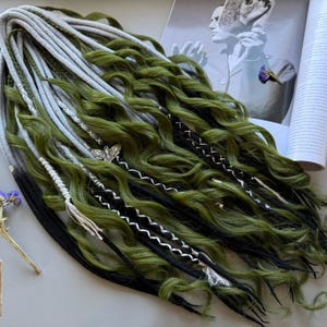May include: Handmade dreadlocks in shades of green, black, and gray. The dreads are styled with wavy ends and decorated with silver-colored butterfly charms and white thread accents. A magazine and a small illustration are in the background.