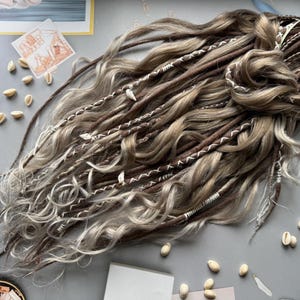 Puede incluir: Conjunto de rastas hechas a mano en tonos marrones y rubios, adornadas con hilo blanco y pequeños acentos de conchas. Las rastas están dispuestas sobre una superficie gris, mostrando su textura y diseño.