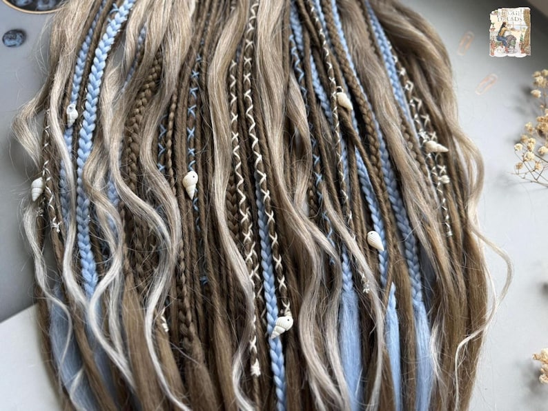 A close-up view of a person's long, braided hairstyle with various colored braids and small white or light blue beads or accessories.