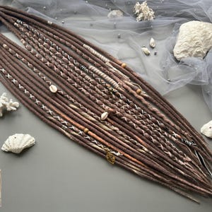 May include: Brown dreadlocks with white and tan accents, adorned with seashells, wooden beads, and leaf charms. The dreads are arranged on a gray surface with decorative coral and a sheer fabric background. The image includes a small logo in the bottom left corner.