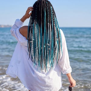 May include: A person with long, black dreadlocks with teal and white accents, standing on a beach. The person is wearing a white, oversized button-down shirt and holding sunglasses. The ocean and sky are visible in the background.
