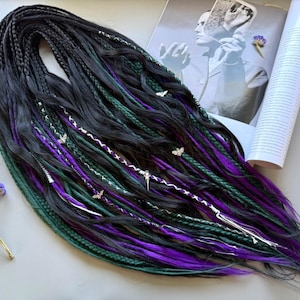 Puede incluir: Dreadlocks negros, verdes y morados con detalles plateados. Los dreadlocks presentan una mezcla de colores y texturas, incluyendo mechones negros, verdes y morados vibrantes. Se incorporan dijes de mariposa y punta plateados.