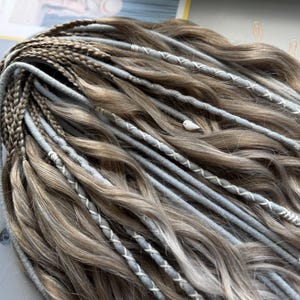 May include: Close-up of a set of brown and gray dreadlocks with light gray accents. The dreadlocks are adorned with white stitching and small decorative bird charms. The hair is styled in a wavy pattern.