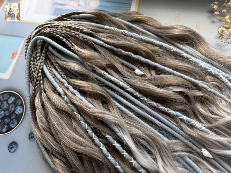 May include: Close-up of a set of brown and gray dreadlocks with light gray accents. The dreadlocks are adorned with white stitching and small decorative bird charms. The hair is styled in a wavy pattern.