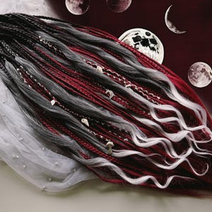 May include: A set of synthetic dreadlocks in shades of black, gray, red, and white, adorned with decorative charms. The dreads are styled with wavy ends and are arranged on a white fabric with small pearls. The background features celestial imagery.