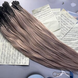 May include: A bundle of ombre hair extensions, transitioning from black to a light brown shade. The extensions are laid out on a surface with sheet music and a pair of scissors.