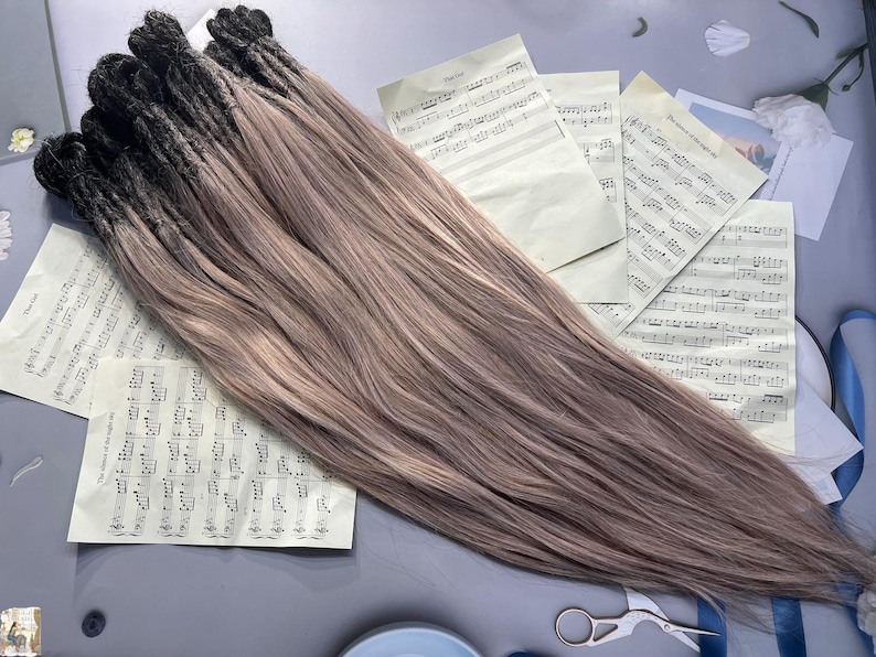 May include: A bundle of ombre hair extensions, transitioning from black to a light brown shade. The extensions are laid out on a surface with sheet music and a pair of scissors.