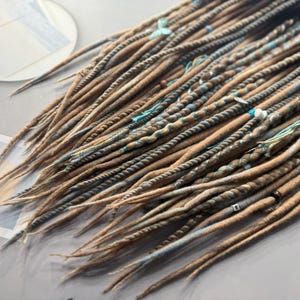 May include: A collection of brown and tan dreadlocks with blue and white accents. The dreads are arranged in a layered pattern, with some sections twisted and others straight. The ends are tapered, and the overall effect is natural and textured.