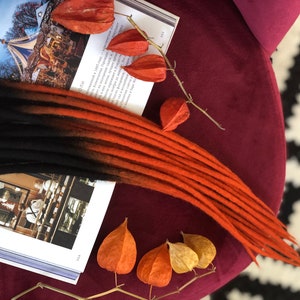 May include: A set of black and orange ombre synthetic dreadlocks. The dreadlocks are arranged on a book with dried orange flowers.