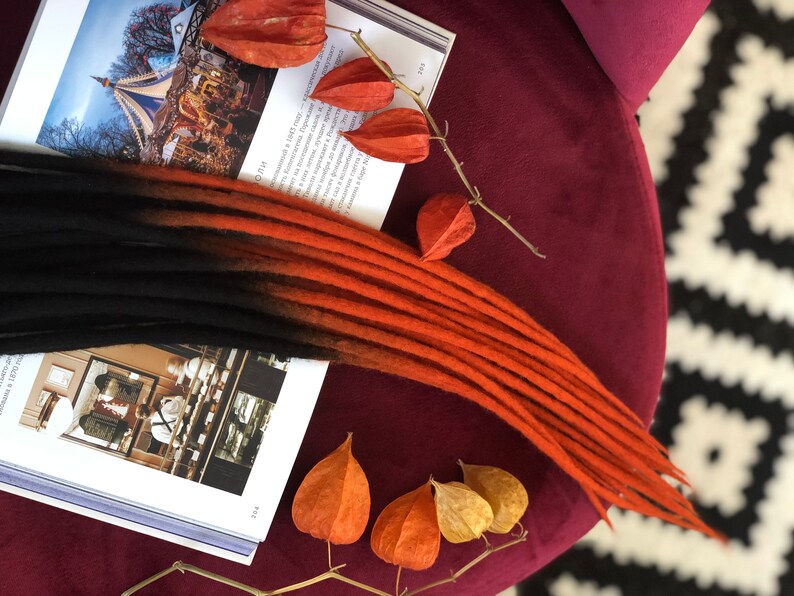 May include: A set of black and orange ombre synthetic dreadlocks. The dreadlocks are arranged on a book with dried orange flowers.