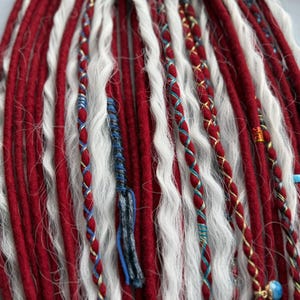 Könnte beinhalten: Nahaufnahme von roten und weißen Dreadlocks. Strähnen von weißem, welligem Haar sind zwischen den roten Dreadlocks. Einige Dreadlocks sind mit blauem und goldenem Faden und kleinen Perlen verziert. Ein auffälliges Haaraccessoire.