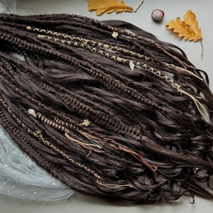 May include: Dark brown dreadlocks with decorative accents. The dreads are styled with various textures and adorned with light-colored string, small charms, and leaf-shaped embellishments. The arrangement is set against a neutral background with autumn leaves and chestnuts.
