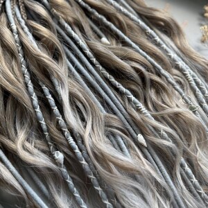 May include: Close-up of a set of handmade dreadlocks in shades of gray and light brown. The dreads are adorned with white thread accents and small decorative beads. The hair is styled with a mix of textures and colors.