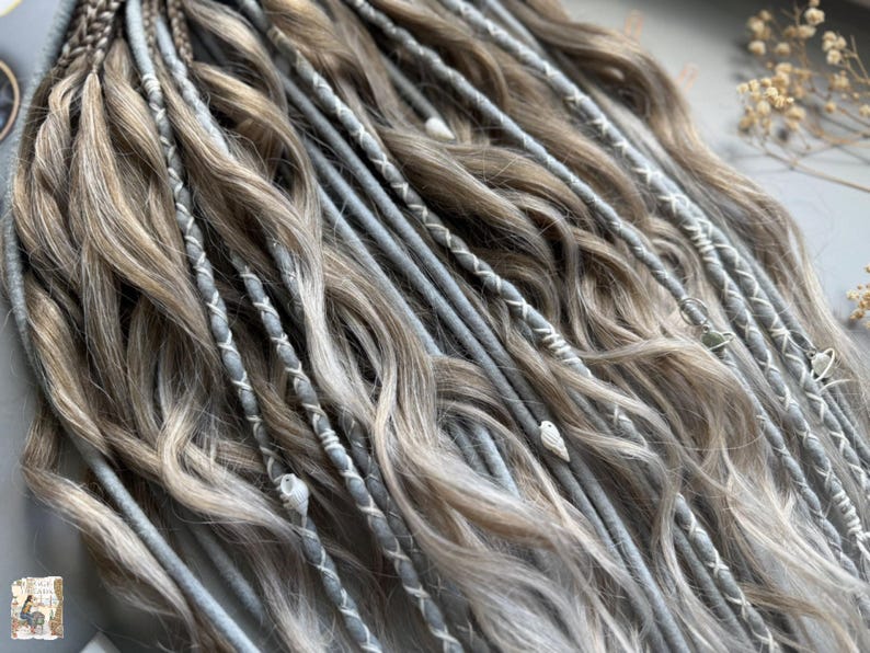May include: Close-up of a set of handmade dreadlocks in shades of gray and light brown. The dreads are adorned with white thread accents and small decorative beads. The hair is styled with a mix of textures and colors.