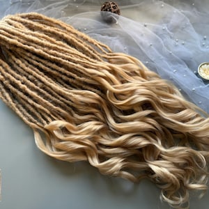May include: Blonde dreadlocks with wavy ends. The dreadlocks are a light brown color at the roots, transitioning to a lighter blonde at the ends. The hair is styled to lay flat and is suitable for a variety of hairstyles.
