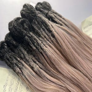May include: A bundle of dreadlocks with a color gradient. The roots are dark, transitioning to a light brown at the ends. The dreadlocks are neatly bundled together, suggesting they are extensions or a hair accessory.