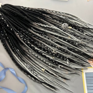 May include: A collection of black, grey, and white synthetic dreadlocks. The dreads are styled with decorative silver accents and small charms. The hair extensions are arranged on a light grey surface, with a blue ribbon and floral accents.
