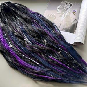 May include: Handmade dreadlocks in shades of black, navy blue, and vibrant purple. The dreads are adorned with silver-colored charms and white accents. A magazine and a small logo are visible in the background.