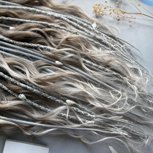 May include: Close-up of a collection of gray and light brown dreadlocks. The dreadlocks are adorned with white thread and small decorative elements. The hair has a wavy texture and a natural, blended color.