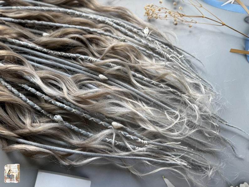 May include: Close-up of a collection of gray and light brown dreadlocks. The dreadlocks are adorned with white thread and small decorative elements. The hair has a wavy texture and a natural, blended color.