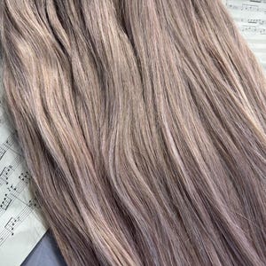 May include: Close-up of bundles of light brown and lavender-toned hair extensions. The hair is straight and has a natural look. The extensions are lying on top of a sheet of vintage sheet music.