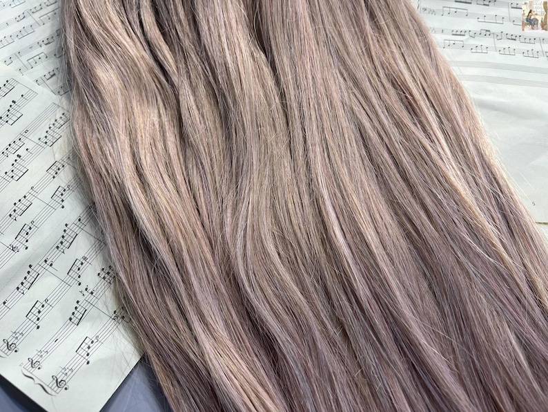 May include: Close-up of bundles of light brown and lavender-toned hair extensions. The hair is straight and has a natural look. The extensions are lying on top of a sheet of vintage sheet music.