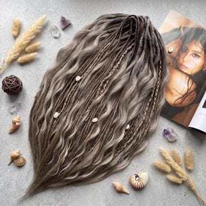 May include: A set of wavy synthetic hair extensions in shades of ash brown and blonde, featuring integrated dreadlocks, braided sections, and small white cowrie shell accents. The hair is arranged in a cascading, layered style with varied textures.