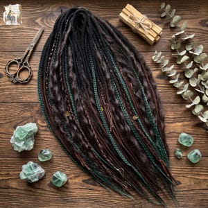 May include: A set of synthetic dreadlocks in shades of black, dark brown, and forest green, featuring braided sections, loose wavy strands, and small gold-toned leaf charms, arranged in a fan shape on a dark wooden surface.