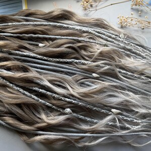 May include: A collection of handmade dreadlocks in shades of brown, gray, and white. The dreads are adorned with white stitching and small decorative elements. The hair extensions are styled in a wavy pattern, creating a natural and textured look.