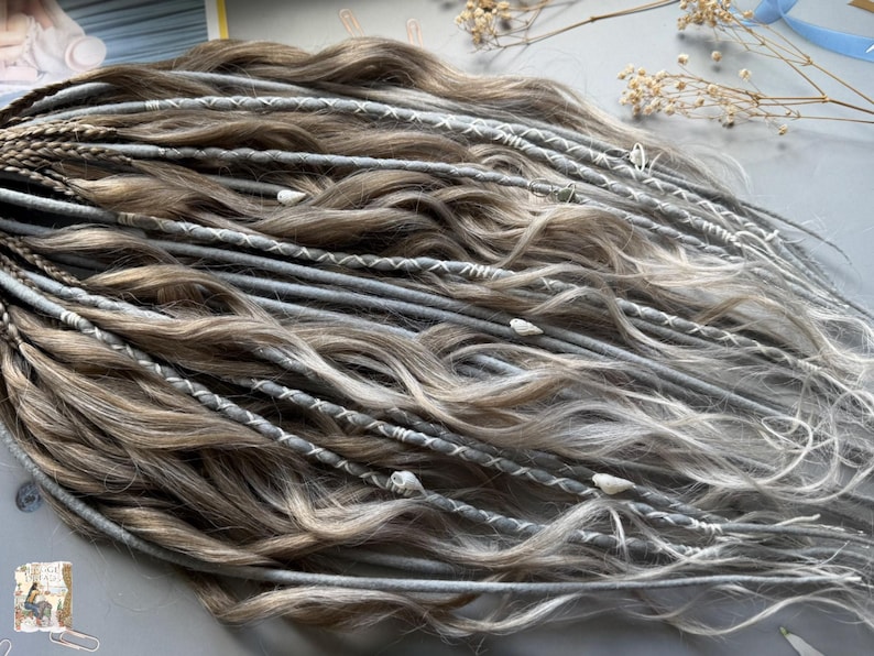 May include: A collection of handmade dreadlocks in shades of brown, gray, and white. The dreads are adorned with white stitching and small decorative elements. The hair extensions are styled in a wavy pattern, creating a natural and textured look.