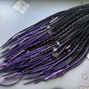 Puede incluir: Un conjunto de rastas sintéticas negras y moradas con secciones trenzadas, dijes plateados en forma de murciélago, calavera y gato, y finos detalles de cinta morada. Las rastas están atadas en la parte superior con una cinta de color morado.
