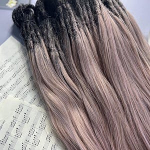 May include: A bundle of dreadlocks with a dark to light purple ombre effect. The dreadlocks are arranged on a surface with sheet music and a white flower.
