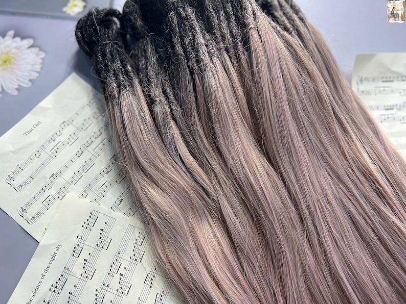 May include: A bundle of dreadlocks with a dark to light purple ombre effect. The dreadlocks are arranged on a surface with sheet music and a white flower.