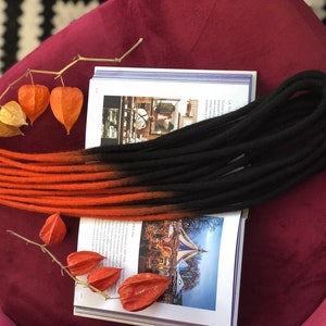 May include: A set of ombre dreadlocks with a black to orange colour gradient. The dreadlocks are made of synthetic hair and are approximately 24 inches long.