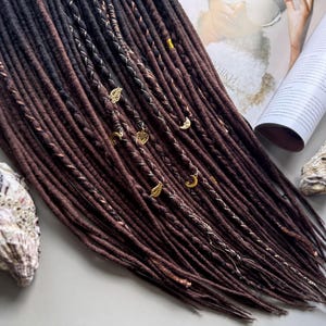 a close-up view of a collection of brown dreadlocks, some adorned with small gold or silver charms, arranged on a surface alongside a magazine and a shell.