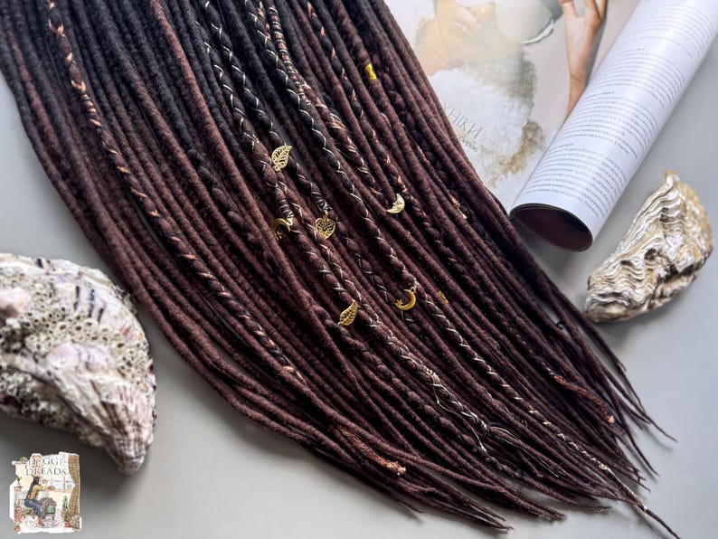 a close-up view of a collection of brown dreadlocks, some adorned with small gold or silver charms, arranged on a surface alongside a magazine and a shell.