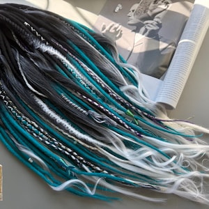 Könnte beinhalten: Eine Sammlung synthetischer Dreadlocks in Schwarz, Grau, Türkis und Weiß. Die Dreads sind in einem fließenden Muster angeordnet, wobei einige Strähnen mit dekorativen Wicklungen verziert sind. Ein Magazin und ein kleines Logo sind im Hintergrund zu sehen.