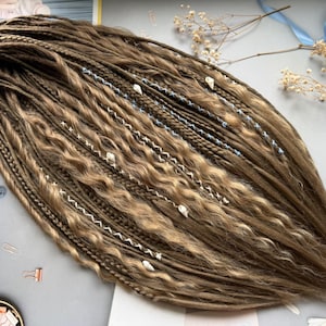 Puede incluir: Dreadlocks marrones y rubios con secciones onduladas y trenzadas. Los dreadlocks están adornados con detalles de hilo blanco y azul y pequeñas decoraciones en forma de concha. Un accesorio para el cabello elegante.