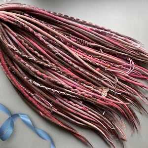 May include: A collection of brown, pink, and white dreadlocks, hand-crafted. The dreadlocks are adorned with white thread, beads, and a silver charm. The dreadlocks are displayed on a grey surface.