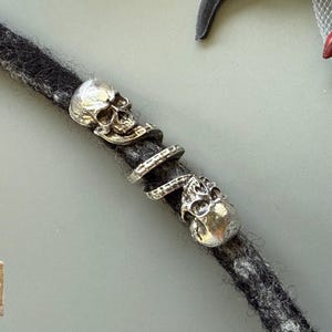 May include: Close-up of a dreadlock with silver skull and ring accents. The dreadlock is dark gray and textured. Two skull charms and several ring charms are visible. The background is a light gray.