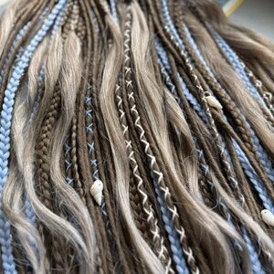 a close-up view of a collection of braided hair strands in various shades of brown and blonde, with small white shells or beads interspersed throughout the braids.