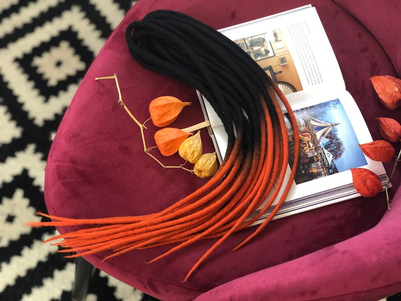 May include: A set of ombre synthetic dreadlocks in black and orange colours. The dreadlocks are arranged on a burgundy surface.