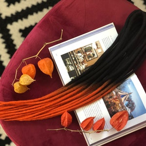 May include: A set of black and orange ombre synthetic dreadlocks. The dreadlocks are laid out on a burgundy surface with a book and dried flower pods.