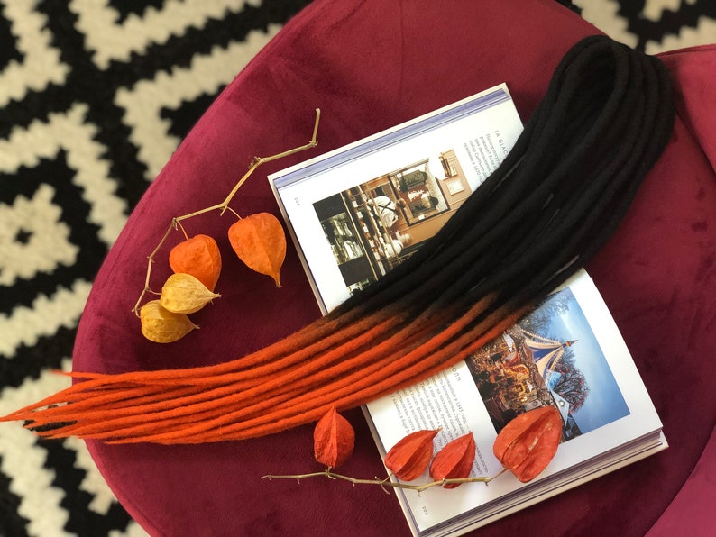May include: A set of black and orange ombre synthetic dreadlocks. The dreadlocks are laid out on a burgundy surface with a book and dried flower pods.