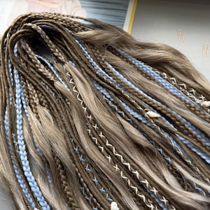 A close-up view of a large number of braided hair strands in various shades of brown and blue, with small white beads or pearls attached to some of the braids.