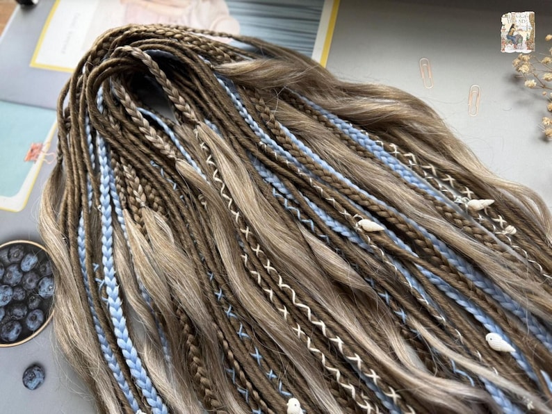 A close-up view of a large number of braided hair strands in various shades of brown and blue, with small white beads or pearls attached to some of the braids.
